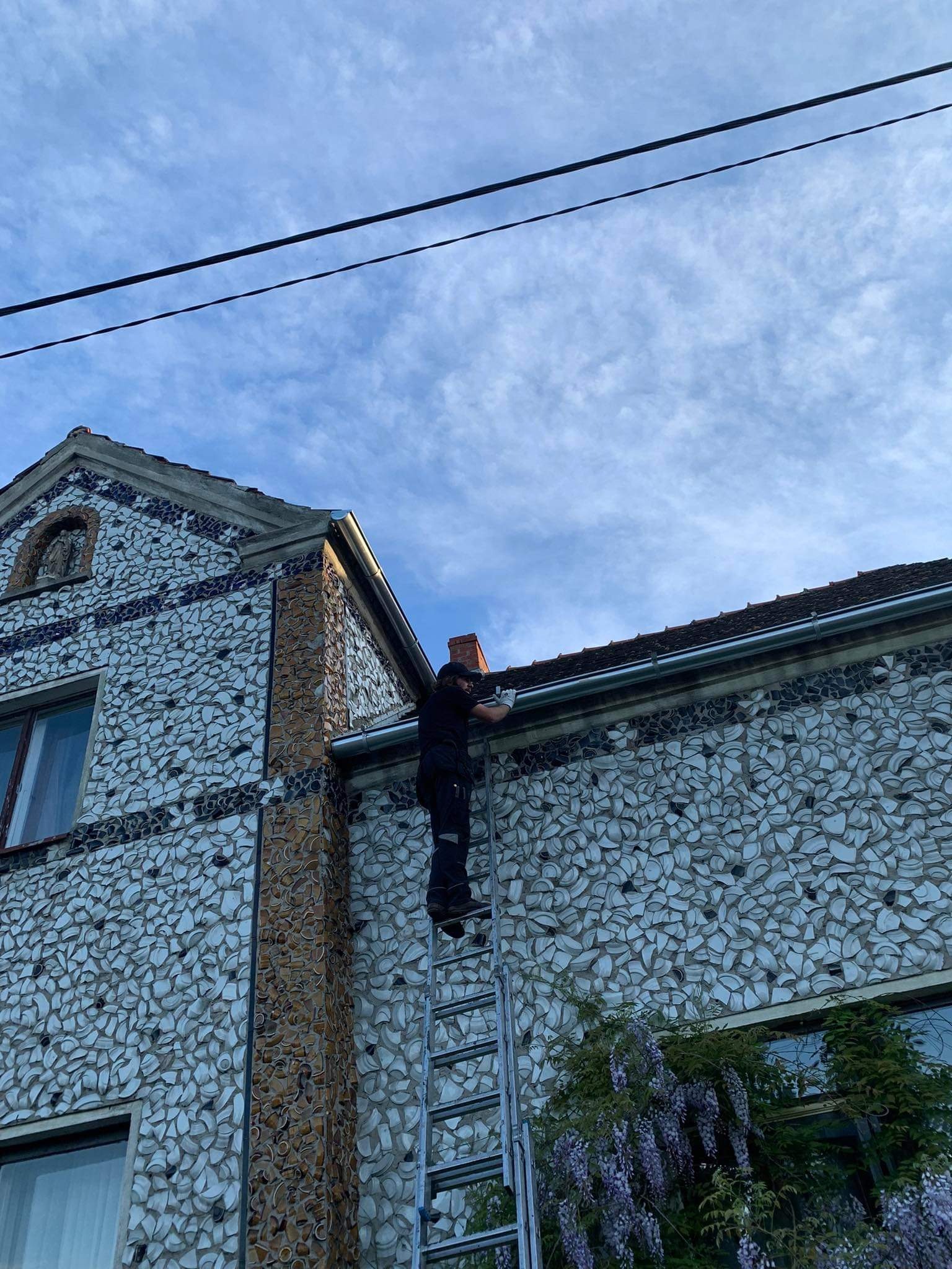 Pracownik na drabinie przy domu z elewacją z potłuczonych talerzy, montujący lub naprawiający rynnę pod okapem dachu.
