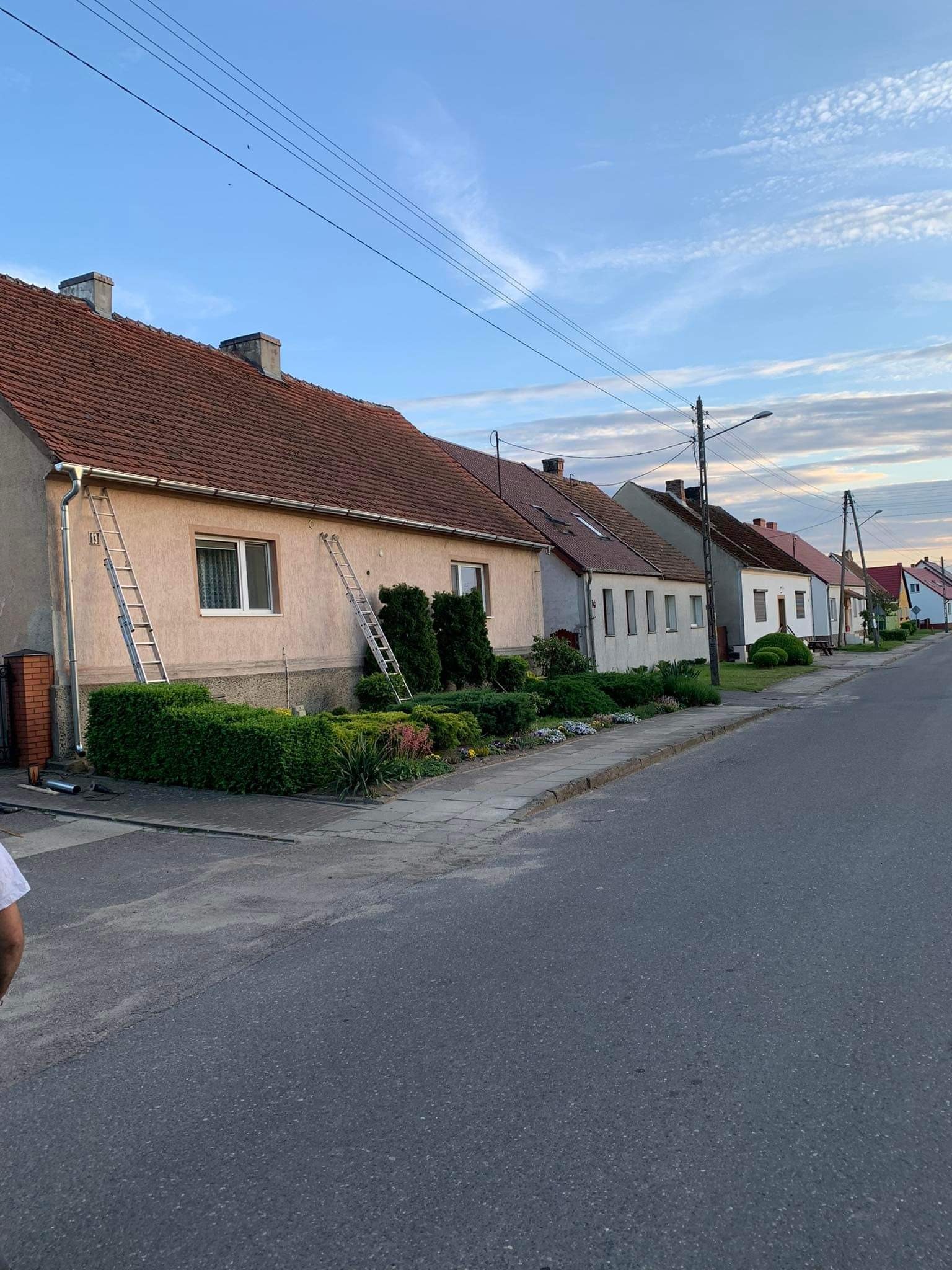 Ulica z rzędem domów jednorodzinnych, dwa metalowe drabiny oparte o ścianę domu z czerwonym dachem, widoczne rynny, krzewy i kwiaty w ogródku przed domem.