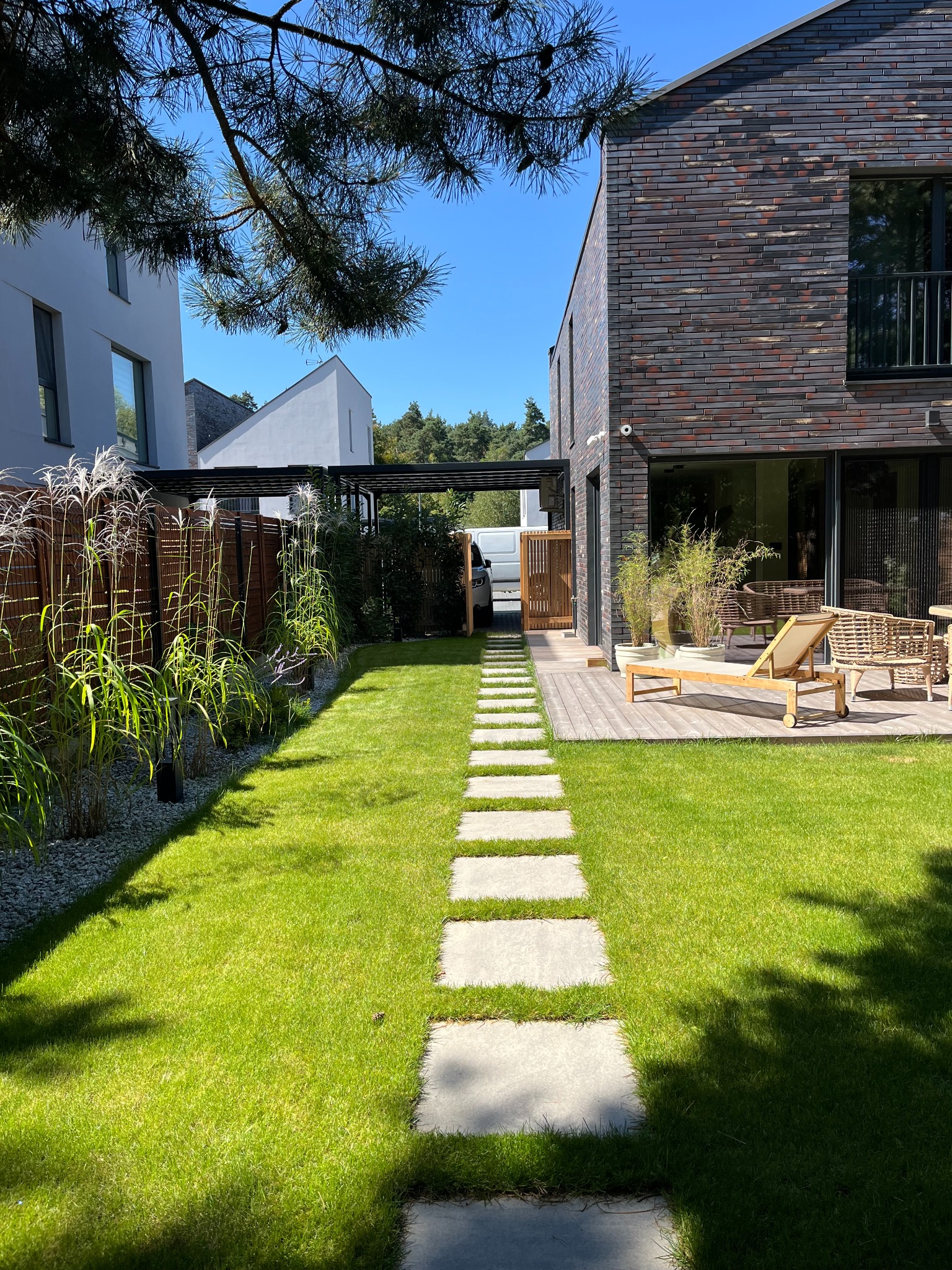 Nowoczesny ogród z ścieżką z płyt, zielonym trawnikiem, drewnianym tarasem z leżakiem i rattanowymi meblami, oraz wysokimi trawami ozdobnymi przy drewnianym ogrodzeniu.