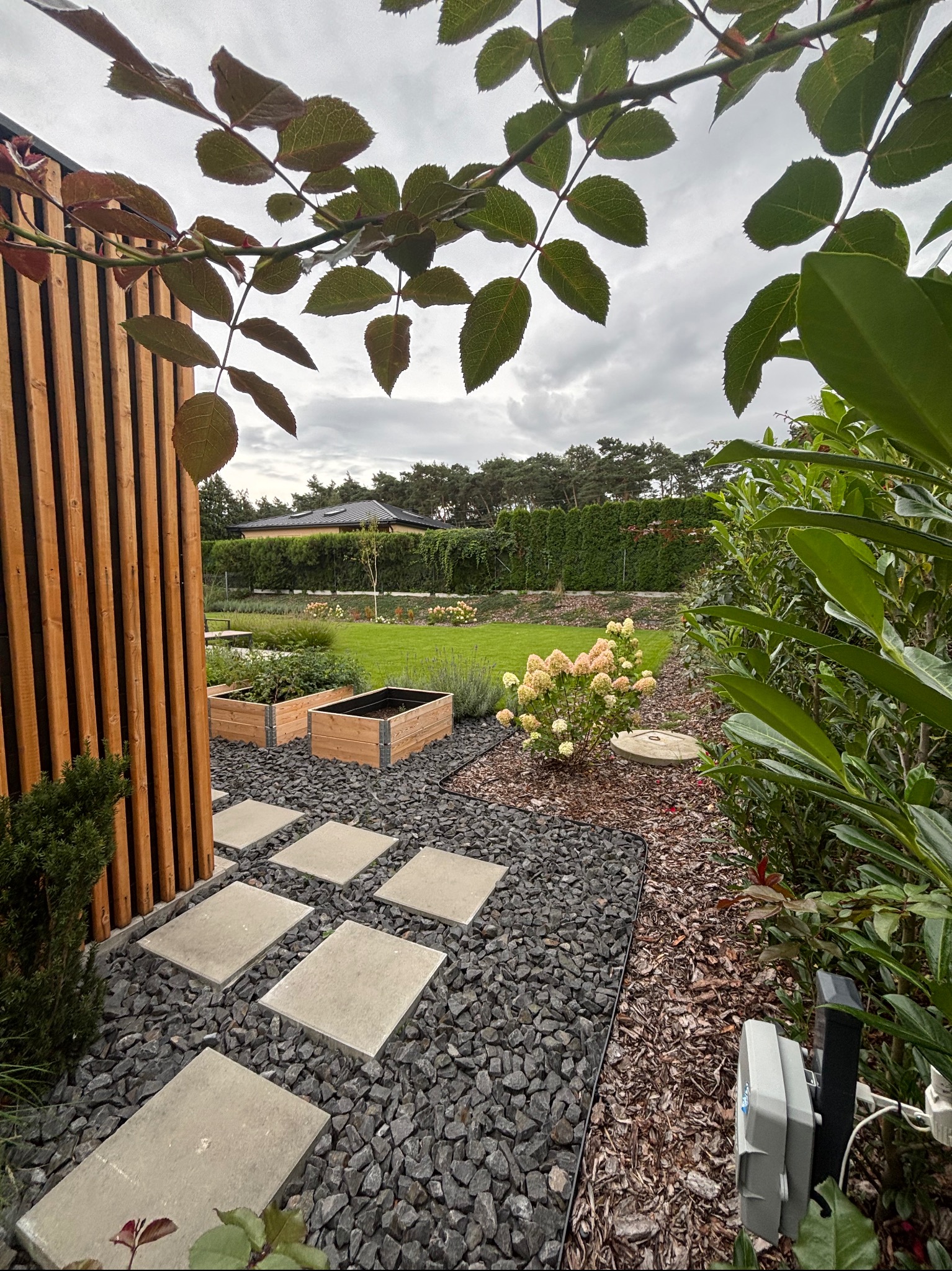 Nowoczesny ogród z kamienną ścieżką, drewnianymi rabatami i bujną zielenią, widziany z perspektywy liści drzewa. Elementy małej architektury wkomponowane w krajobraz.