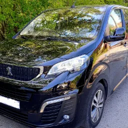 Czarny, lśniący Peugeot Expert z anteną na dachu, zaparkowany na asfaltowej drodze w słoneczny dzień, z widocznym odblaskiem drzew w karoserii.