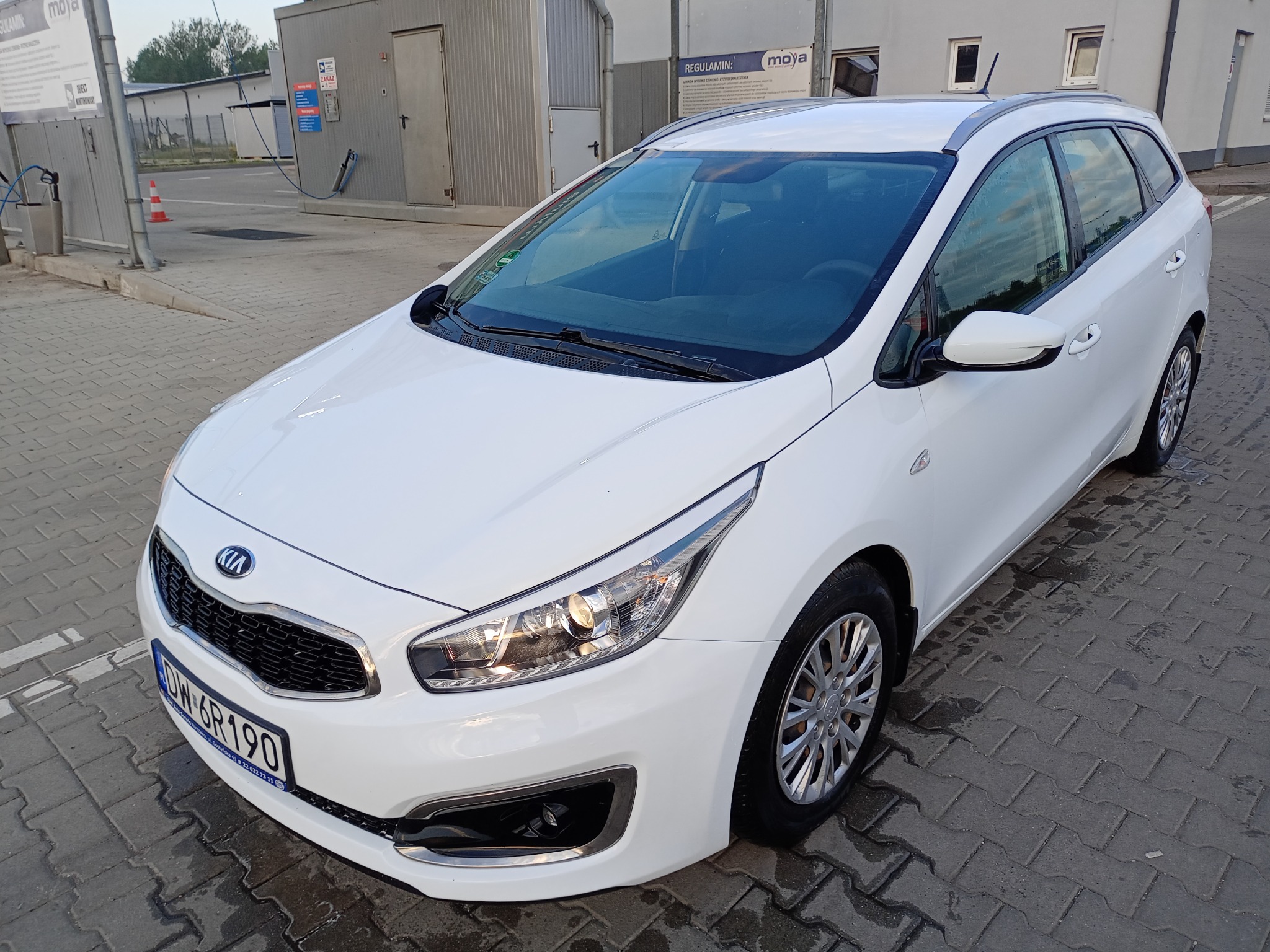 Biała Kia Ceed kombi na kostce brukowej przed myjnią samochodową Moya, widoczne detale karoserii i tablica rejestracyjna.