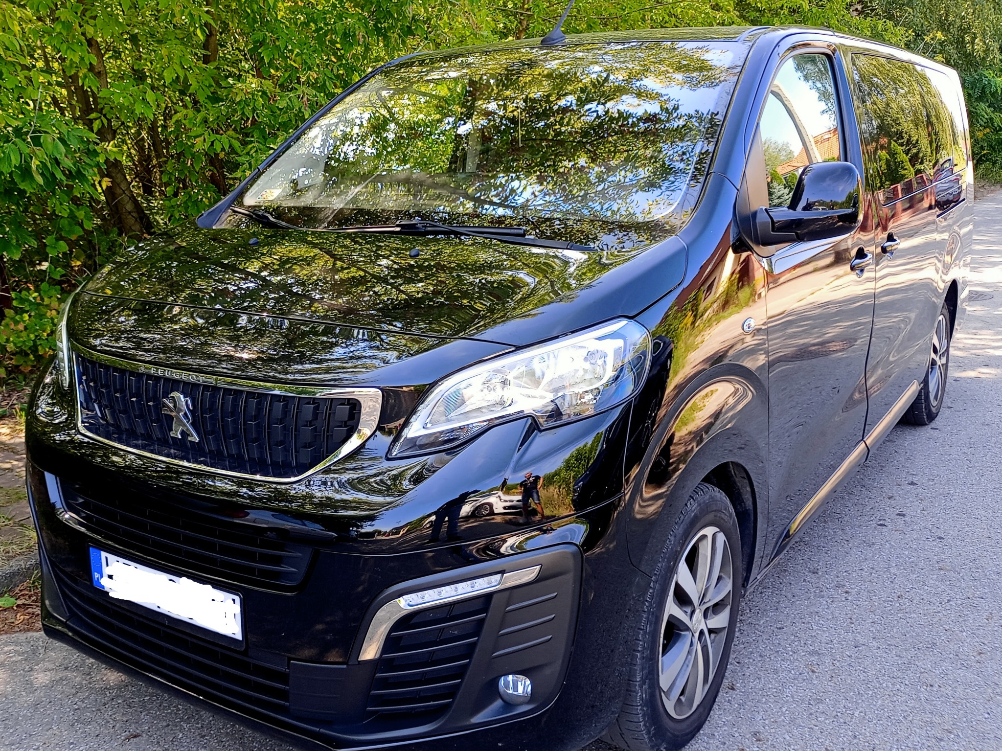 Czarny, lśniący Peugeot Expert z anteną na dachu, zaparkowany na asfaltowej drodze w słoneczny dzień, z widocznym odblaskiem drzew w karoserii.
