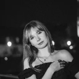 Czarno-białe portretowe ujęcie kobiety w sukience z odkrytymi ramionami, z delikatnym uśmiechem i długimi włosami z grzywką, na rozmytym tle z punktami świetlnymi.