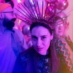 Kobieta z brokatem na policzkach i koroną z zip-ties w neonowym oświetleniu, w tle balony i dwie osoby.