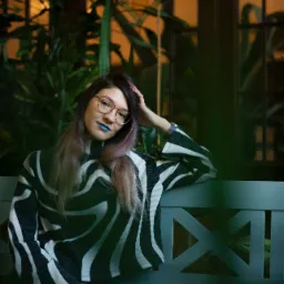 Portret kobiety w okularach z niebieską szminką, siedzącej na ławce z zielonymi roślinami w tle.