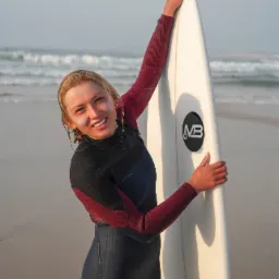 Uśmiechnięta blondynka w piance surfuje, trzymając deskę z logo MB na tle piaszczystej plaży i falującego morza.