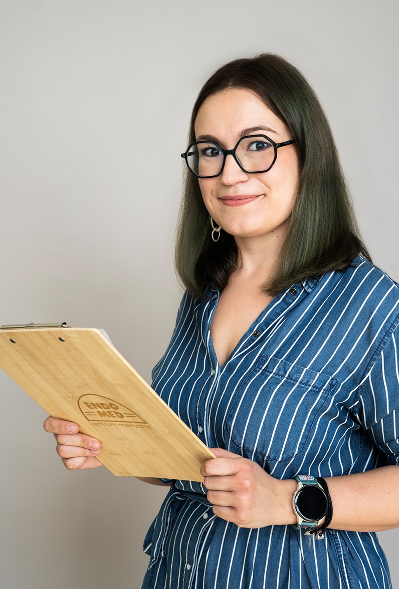 Portret uśmiechniętej kobiety w okularach, trzymającej drewnianą podkładkę z logo Centrum Medycznego ENDOMED, ubrana w niebieską koszulę w paski, z zegarkiem na nadgarstku.