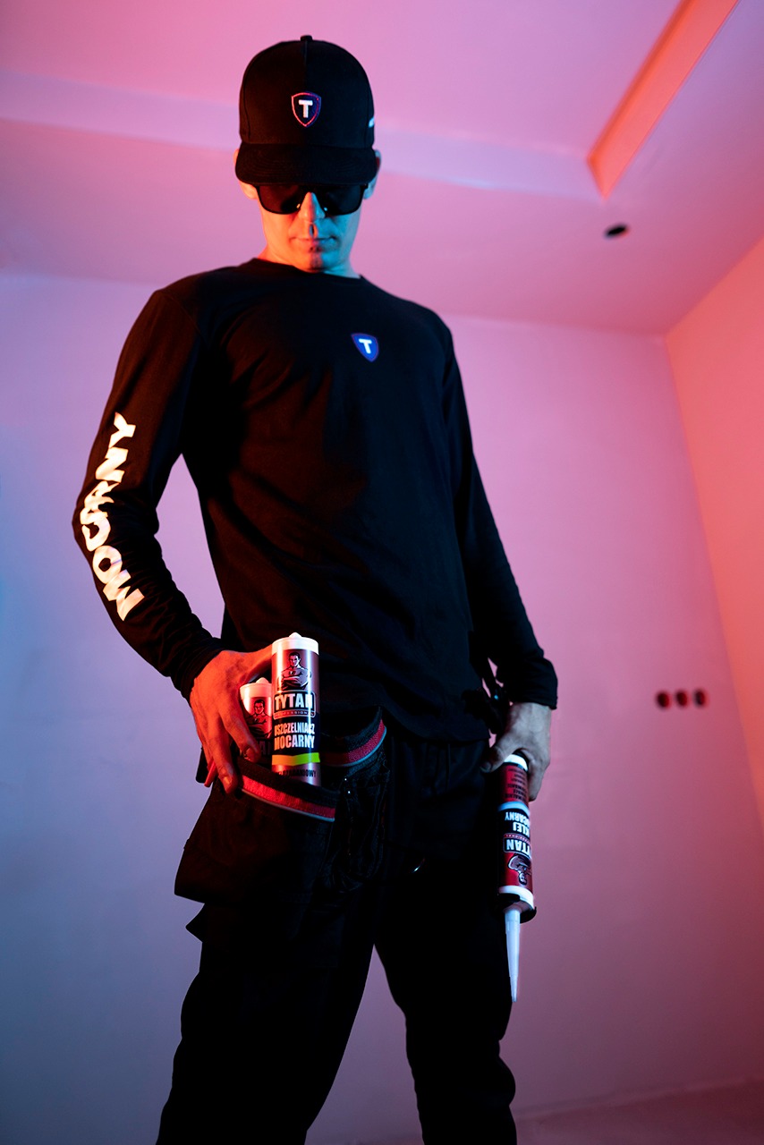 Mężczyzna w czarnym ubraniu i czapce z daszkiem, trzymający tuby uszczelniacza Tytan Mocarny w neonowym oświetleniu, prezentacja produktu.