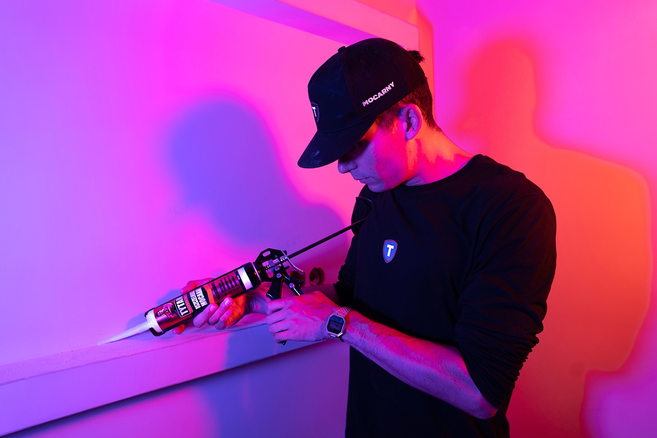 Mężczyzna w czarnej koszulce i czapce z daszkiem, w neonowym oświetleniu, aplikuje silikon za pomocą pistoletu do spoinowania, widoczny zegarek na nadgarstku.