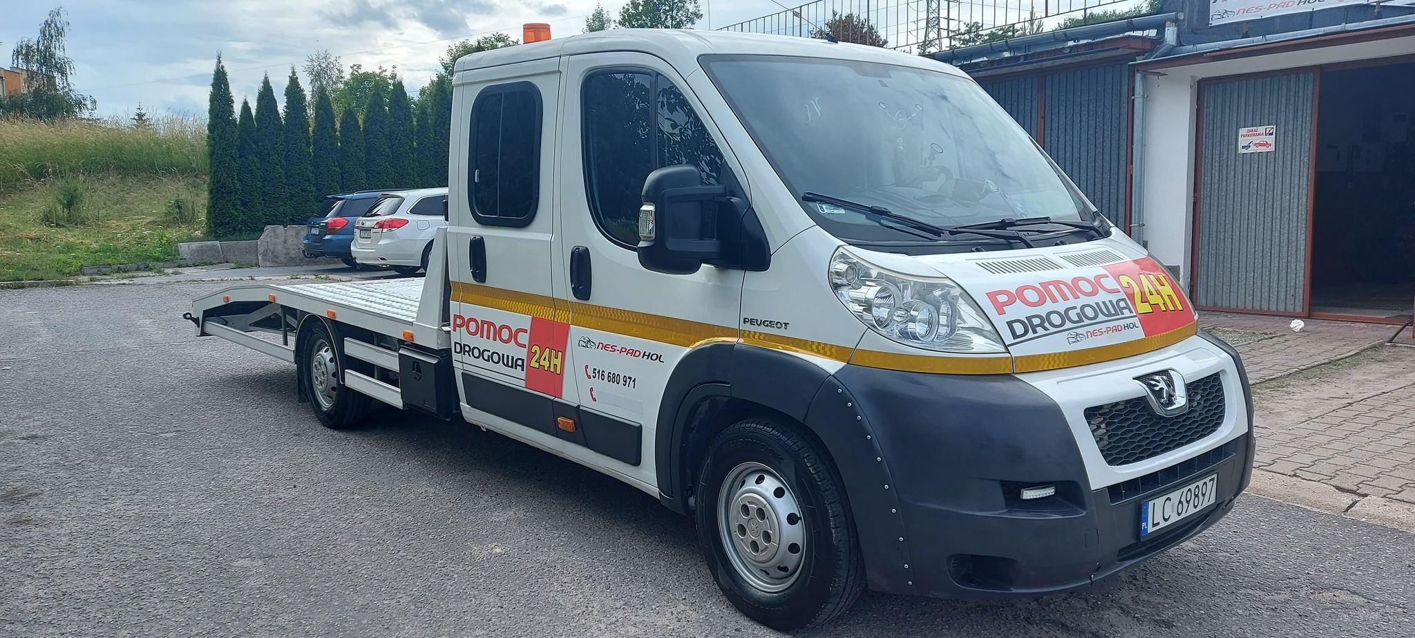 Biała laweta Peugeot z napisem 'Pomoc Drogowa 24H' zaparkowana przed garażem, widok z boku, na tablicy rejestracyjnej widoczny numer LC 69897.
