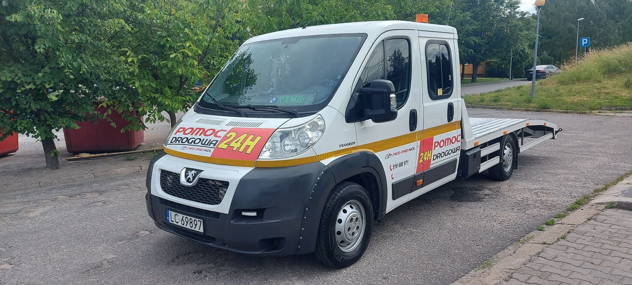 Biała laweta Peugeot z napisem 'Pomoc Drogowa 24H' zaparkowana na chodniku, z zielonymi drzewami i czerwonym kontenerem w tle.