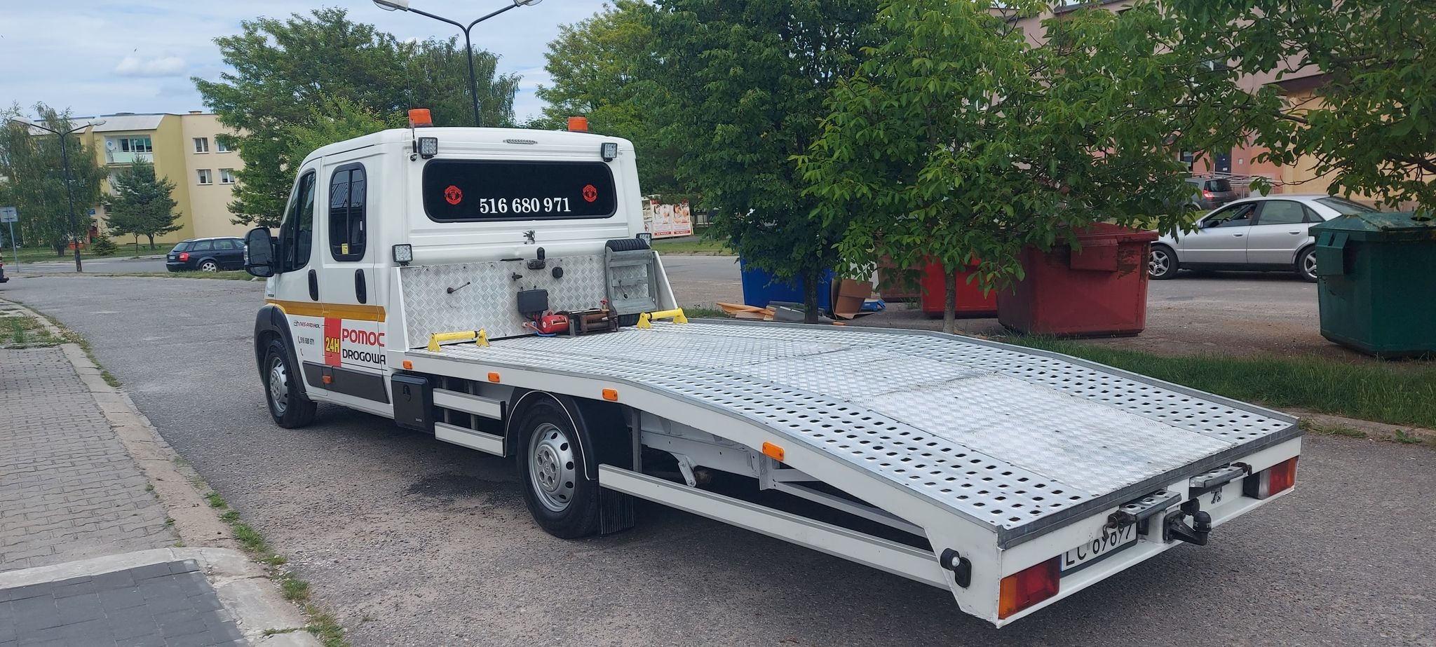 Biała laweta z numerem telefonu w tylnej szybie, zaparkowana na ulicy w dzień.