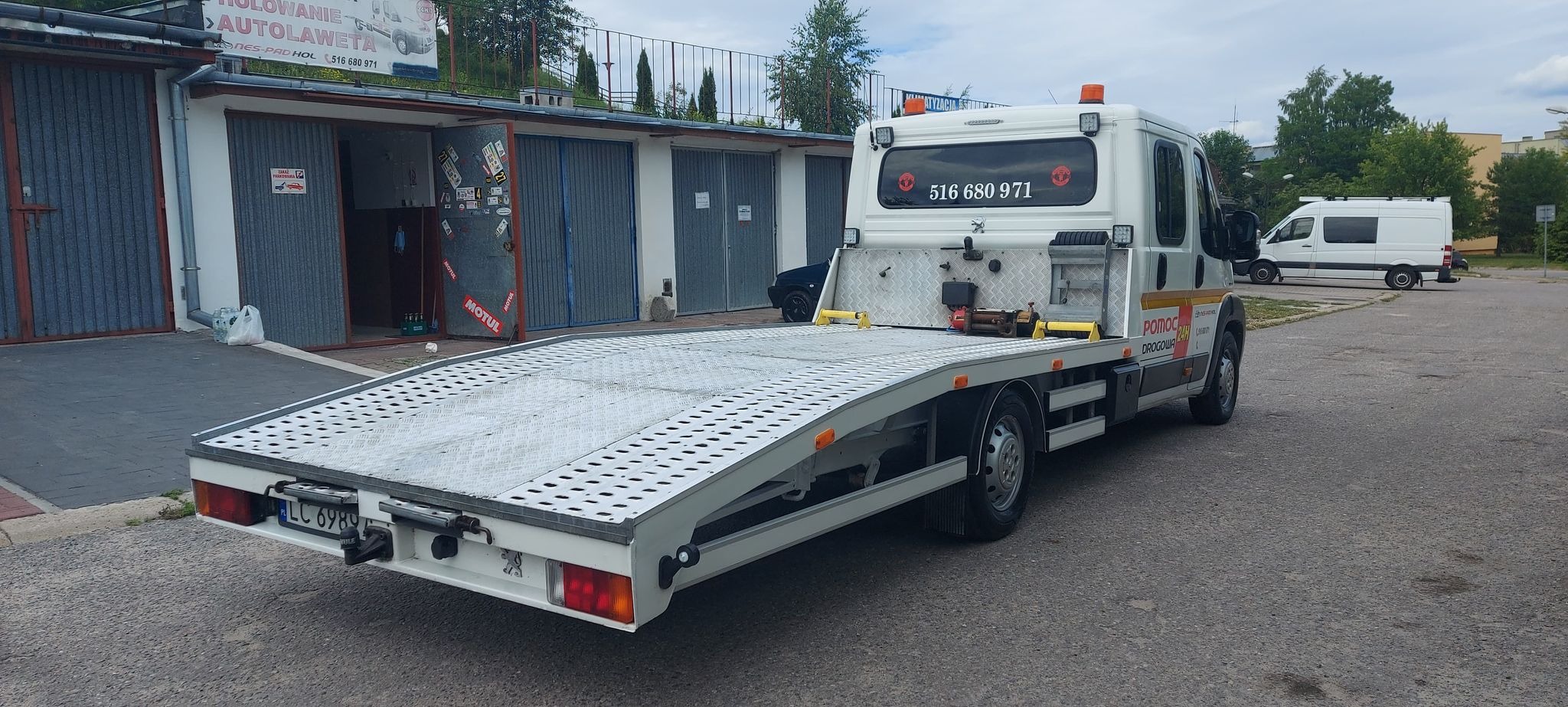 Biała laweta z platformą o metalowej fakturze, zaparkowana przed budynkiem warsztatu z podniesioną platformą, widoczny numer telefonu na kabinie i napis Pomoc Drogowa 24h.