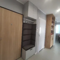 Przedpokój z zabudową: drewniane drzwi wejściowe, szara szafka z wieszakami i tapicerowanym siedziskiem, białe drzwi wewnętrzne, drewniany regał z półkami.