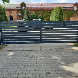 Nowoczesna, przesuwna brama wjazdowa o poziomych panelach w kolorze grafitowym, zamontowana przed budynkiem z czerwonej cegły porośniętym bluszczem. Widoczny fragment chodnika z kostki brukowej...