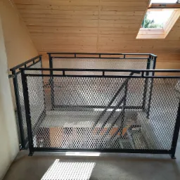 Metalowa balustrada z siatki zabezpieczająca schody w surowym wnętrzu poddasza z drewnianą boazerią i oknem dachowym.