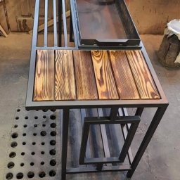 KUE PROJEKT SP. Z O.O. - Metalowa konstrukcja grilla z drewnianym blatem i rusztem, widoczne ślady obróbki, wnętrze warsztatu.