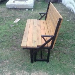 KUE PROJEKT SP. Z O.O. - Drewniana ławka z metalową konstrukcją na trawie, z widocznymi elementami spawania i skręcania, ustawiona obok betonowego elementu i białego płotu.