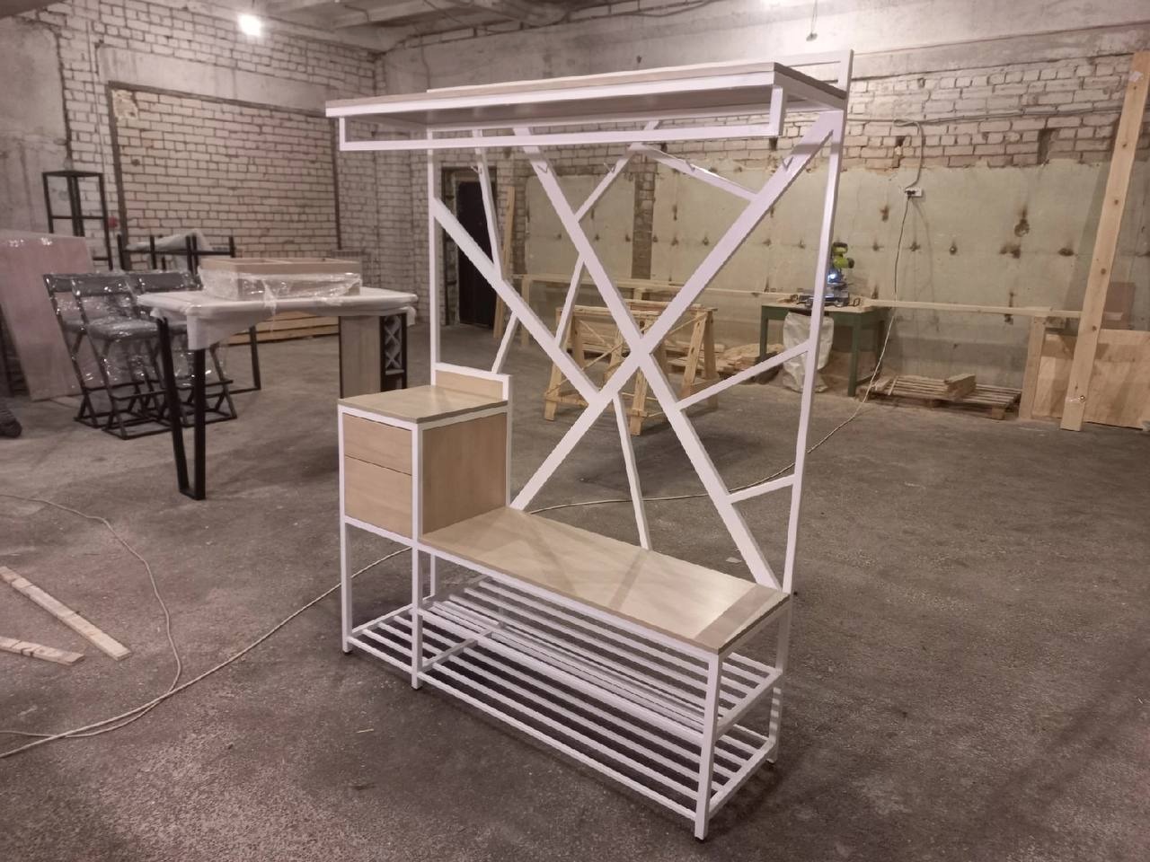 Metalowa konstrukcja w stylu loft z drewnianymi elementami: wieszak na ubrania z półką na buty i szafką, widoczne surowe wnętrze warsztatu z ceglaną ścianą.