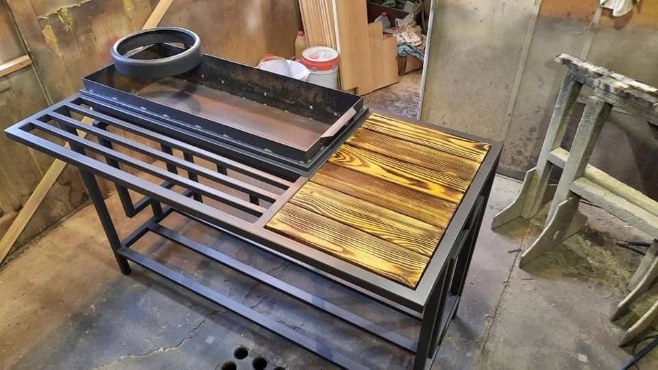 Metalowa konstrukcja z drewnianym blatem, prawdopodobnie stół warsztatowy lub element grilla, w tle widoczne narzędzia i materiały w warsztacie.