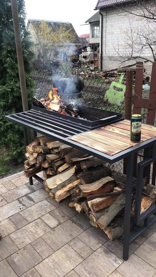 Metalowy grill z paleniskiem, stertą drewna opałowego i piwem na drewnianym blacie, ustawiony na kostce brukowej przed domem z siatką ogrodzeniową w tle.