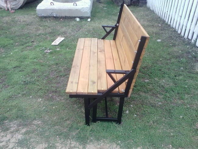 Drewniana ławka z metalową konstrukcją na trawie, z widocznymi elementami spawania i skręcania, ustawiona obok betonowego elementu i białego płotu.