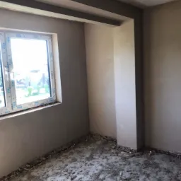 Pokój po nałożeniu gładzi gipsowej, widoczne świeżo otynkowane ściany i zabrudzona podłoga, okno PCV marki VEKA.