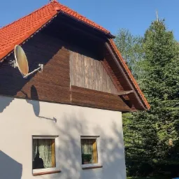 Dwupiętrowy dom z czerwonym dachem z dachówki, brązową drewnianą elewacją na poddaszu, anteną satelitarną i dwoma oknami na parterze, otoczony zielonymi drzewami iglastymi.