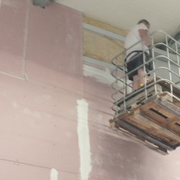 A.L. serwis budowlany - Pracownik na podnośniku koszowym, szpachluje łączenia płyt karton-gips na ścianie wewnątrz budynku, widoczne ocieplenie wełną mineralną w przestrzeni między ścianą a sufitem.