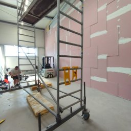 A.L. serwis budowlany - Montaż różowych płyt gipsowo-kartonowych na ścianie, widoczne rusztowanie, poziomice, w tle wózek widłowy i pracownik w trakcie pracy.