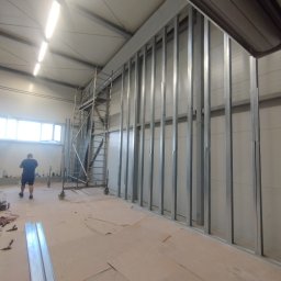 A.L. serwis budowlany - Metalowa konstrukcja szkieletowa ścianki działowej w hali, widoczne profile pionowe i poziome, w tle rusztowanie i pracownik w oddali.