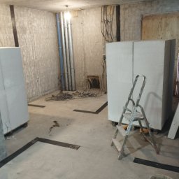 A.L. serwis budowlany - Wnętrze w trakcie prac wykończeniowych: bloki z betonu komórkowego ustawione w surowym pomieszczeniu, widoczne instalacje, drabina aluminiowa i fragmenty taśmy wyznaczającej obszar roboczy.