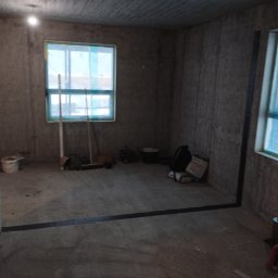 A.L. serwis budowlany - Surowy stan wnętrza domu w trakcie budowy: ściany z betonu komórkowego, okna zabezpieczone folią, widoczne narzędzia i materiały budowlane, brak wykończenia podłogi.