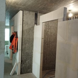 A.L. serwis budowlany - Wnętrze w trakcie budowy z surowymi ścianami z betonu komórkowego, widoczna pomarańczowa kamizelka odblaskowa i czarny plecak powieszone na ścianie, fragment instalacji elektrycznej na suficie.