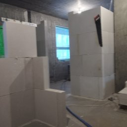 A.L. serwis budowlany - Wznoszenie ścian z białych bloczków, widoczna piła ręczna na ścianie, poziomica i folia ochronna, surowy stan wykończenia wnętrza.