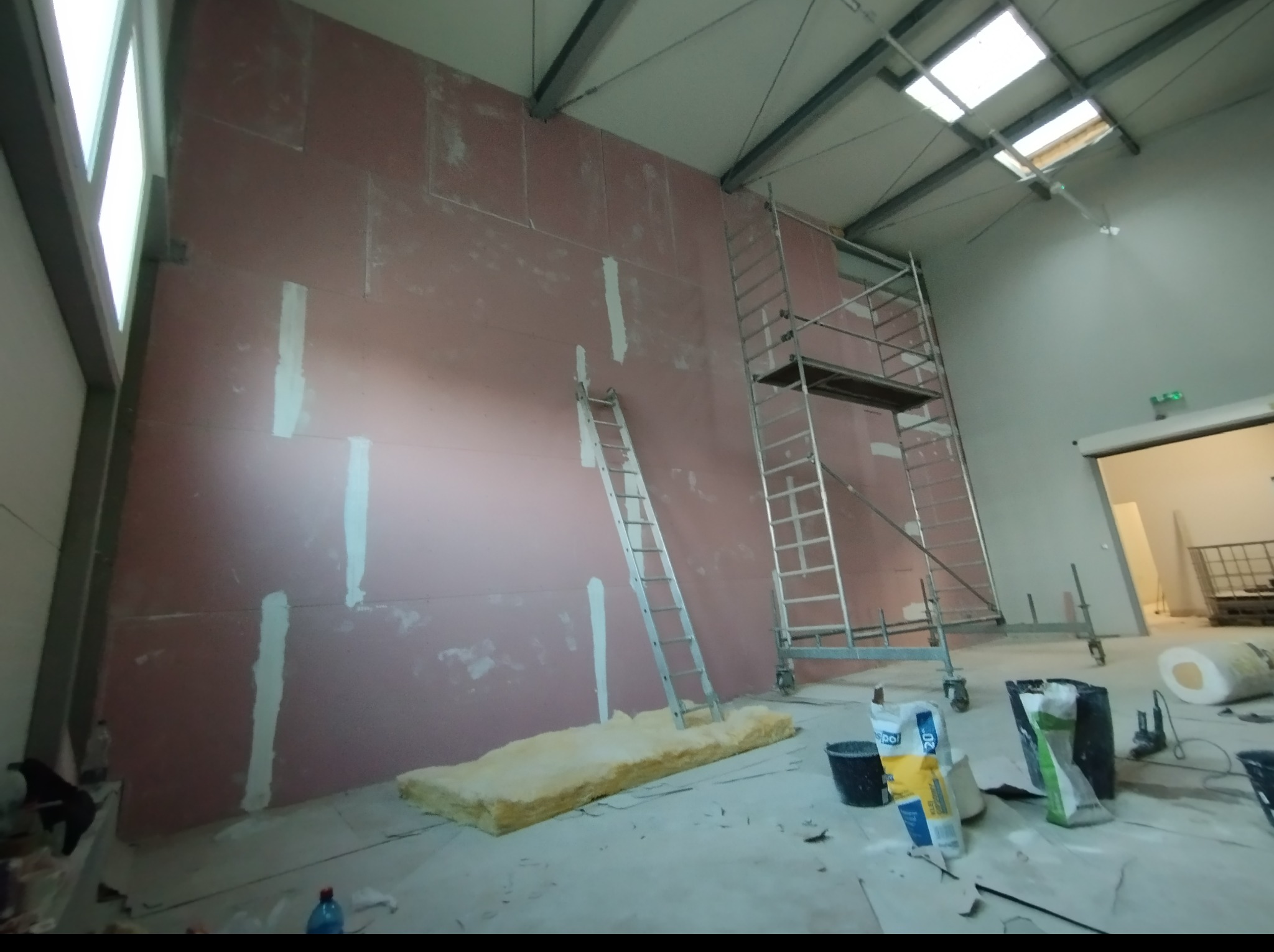 Montaż płyt kartonowo-gipsowych w hali przemysłowej, widoczne rusztowanie, drabina i materiały izolacyjne.