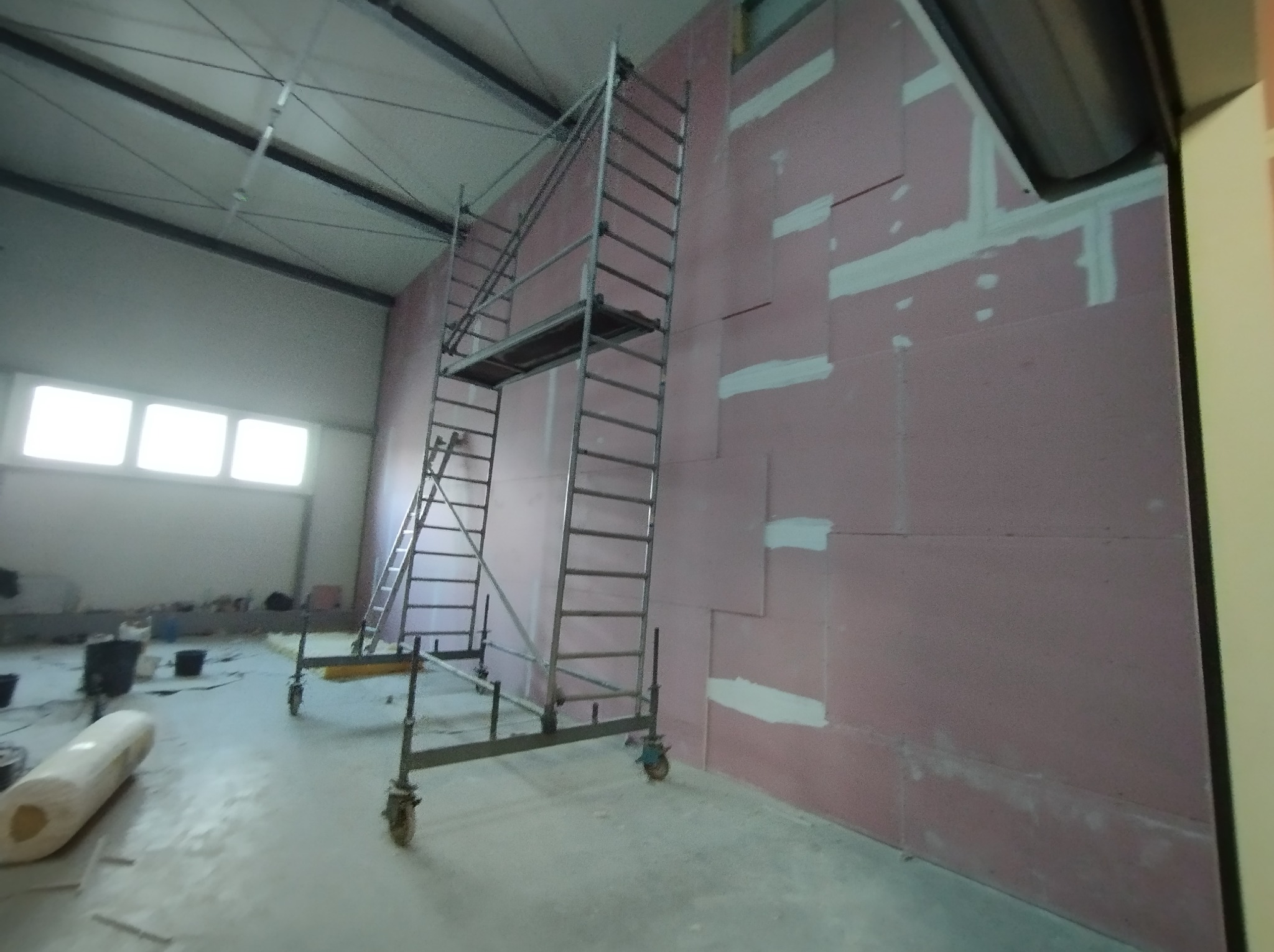 Wnętrze hali w trakcie prac wykończeniowych: ściana z płyt izolacyjnych, częściowo uszczelniona, rusztowanie na kółkach, widoczne worki z materiałem i pojemniki na podłodze.