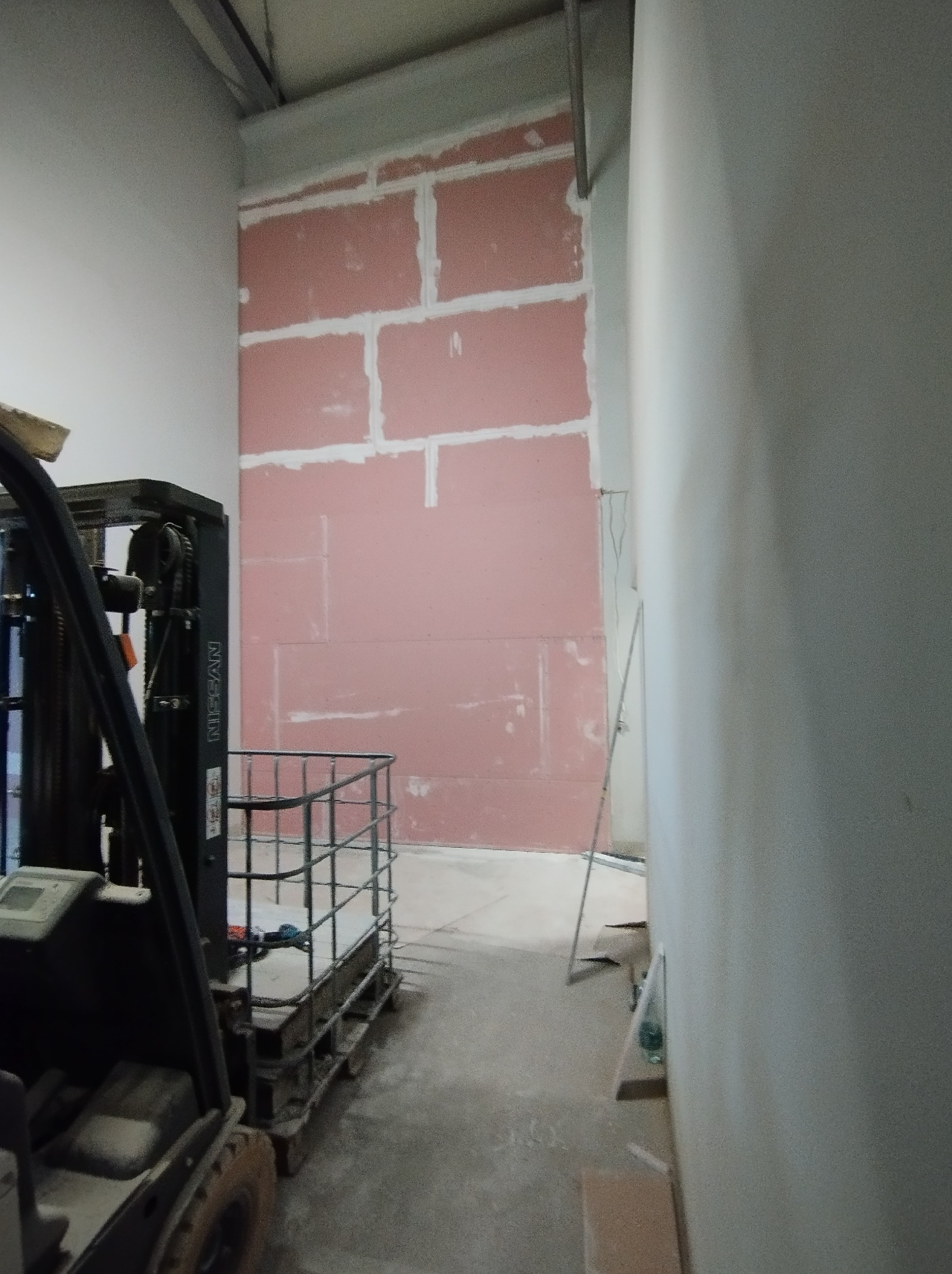 Montaż różowych płyt gipsowo-kartonowych na ścianie, widoczny wózek widłowy i drabina, ślady po szpachlowaniu na łączeniach płyt.