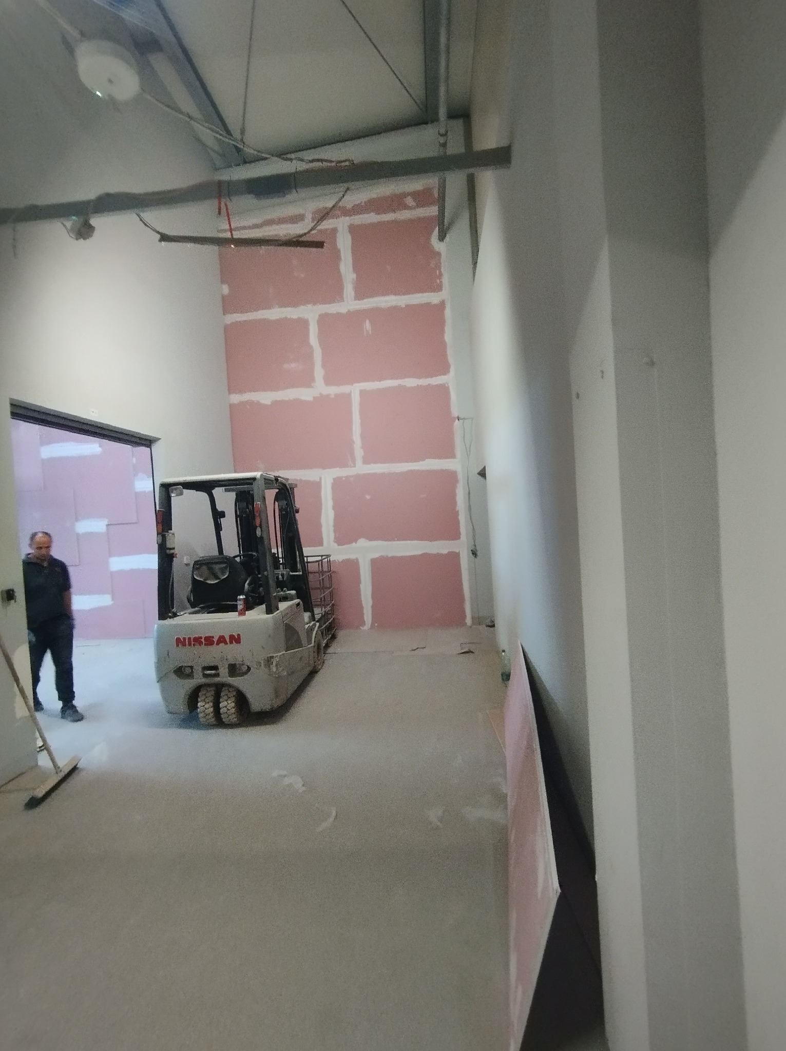 Wnętrze w trakcie montażu płyt kartonowo-gipsowych na ścianie, z widocznym wózkiem widłowym Nissan i mężczyzną z miotłą w tle.