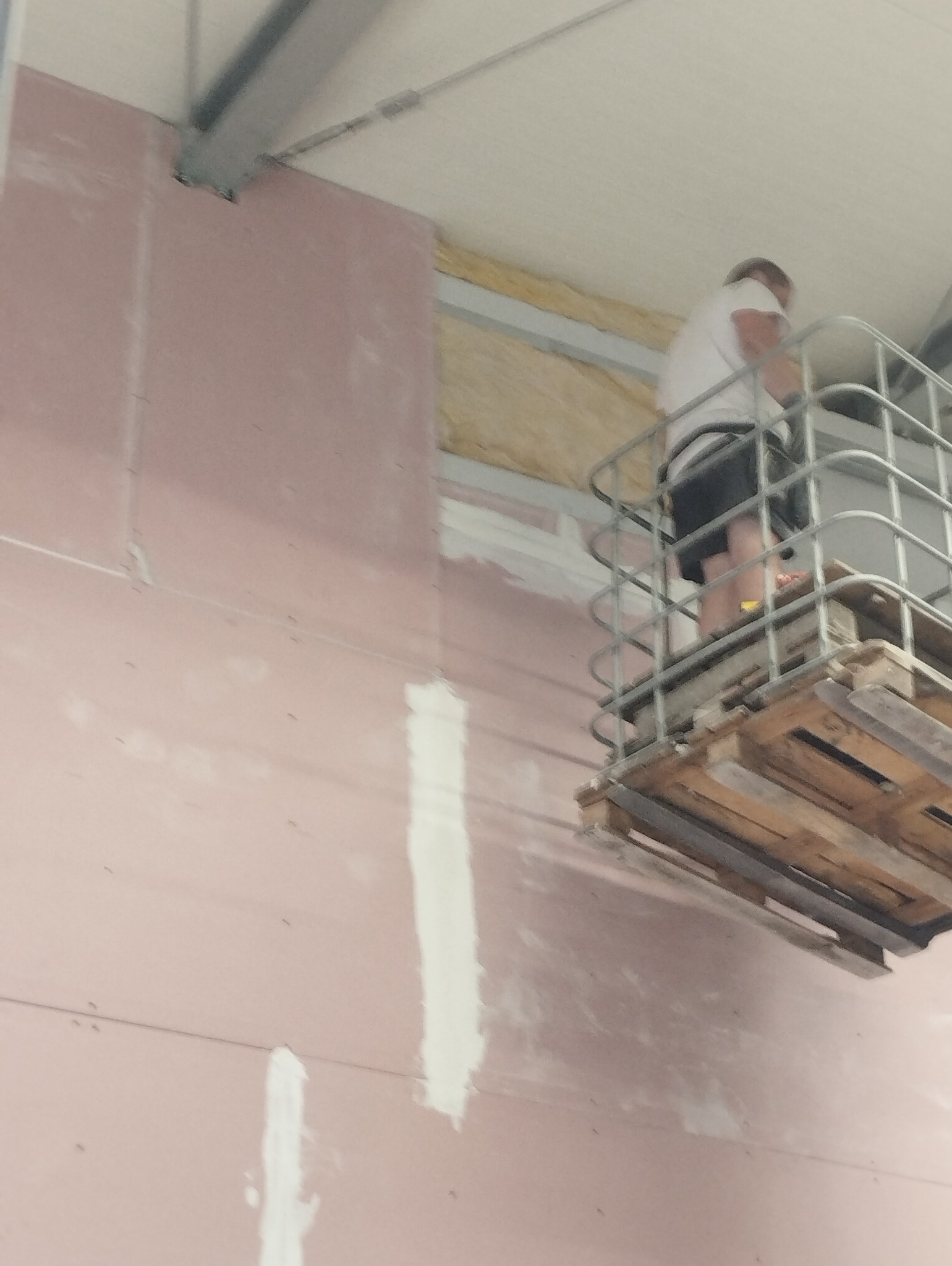 Pracownik na podnośniku koszowym, szpachluje łączenia płyt karton-gips na ścianie wewnątrz budynku, widoczne ocieplenie wełną mineralną w przestrzeni między ścianą a sufitem.