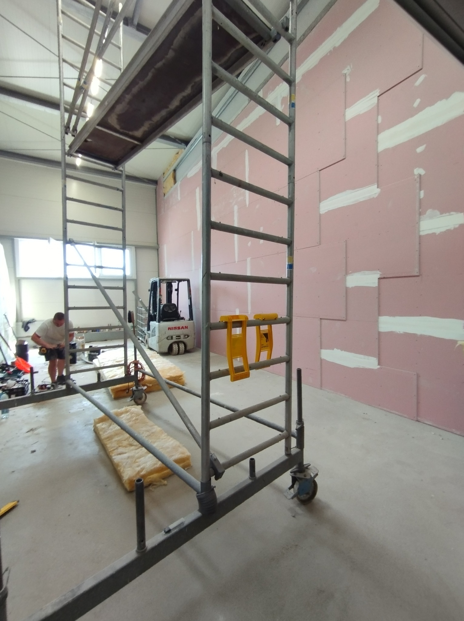 Montaż różowych płyt gipsowo-kartonowych na ścianie, widoczne rusztowanie, poziomice, w tle wózek widłowy i pracownik w trakcie pracy.