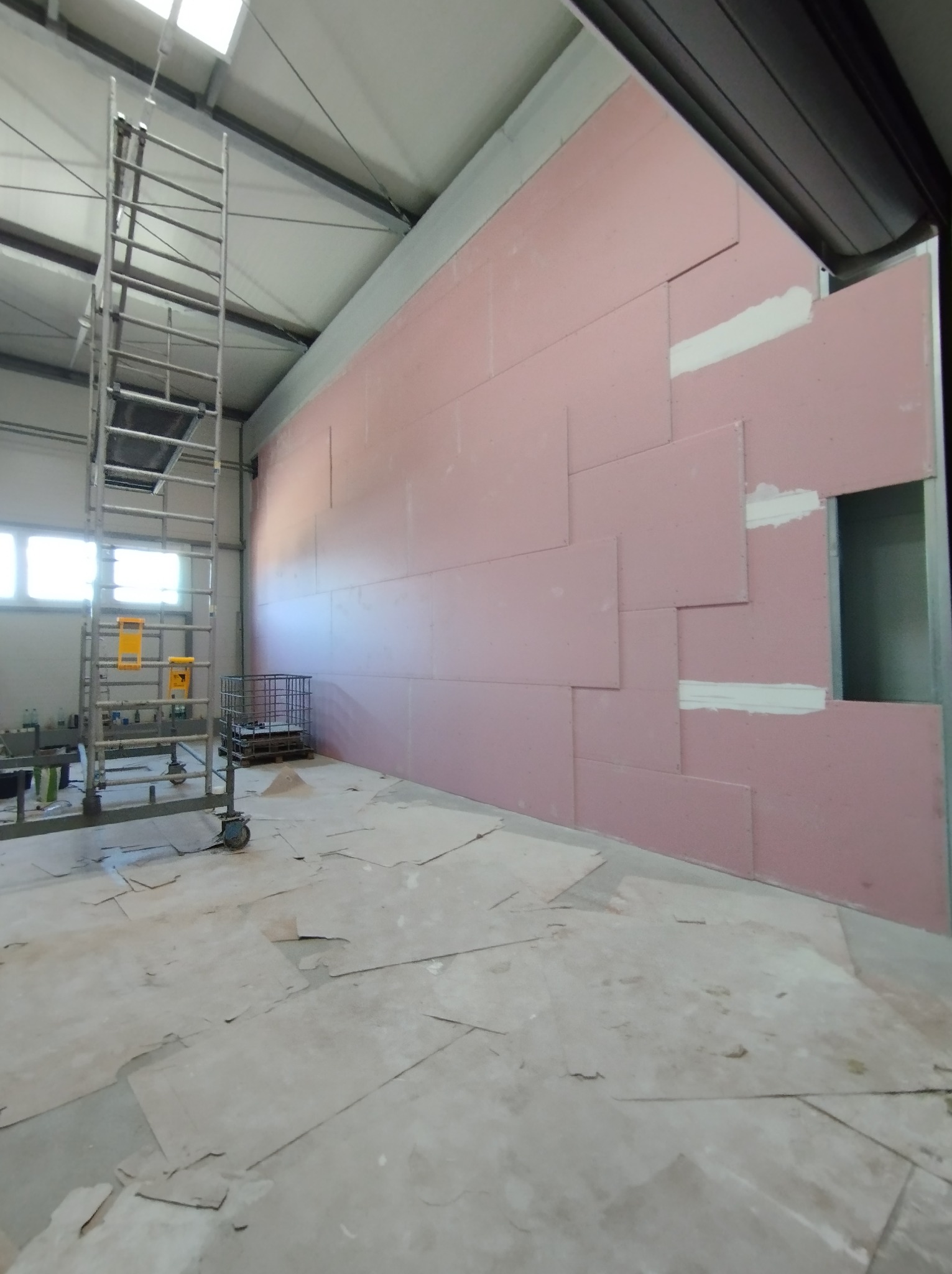Montaż różowych płyt gipsowo-kartonowych na ścianie w hali, widoczne łączenia spoinowane gipsem, metalowe rusztowanie i porozrzucane fragmenty płyt na podłodze.