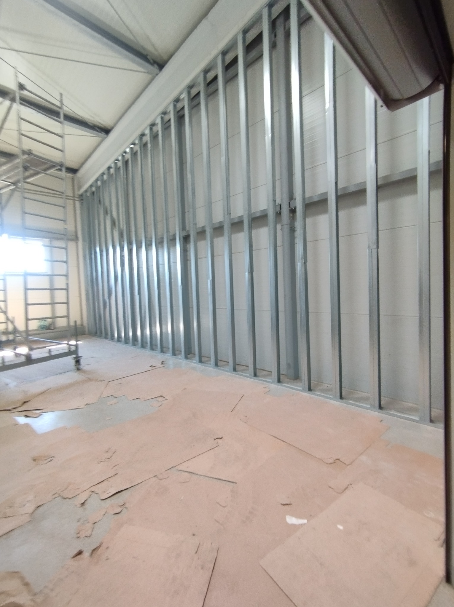 Metalowa konstrukcja szkieletowa ściany w hali przemysłowej, widoczne płyty podłogowe i rusztowanie w tle.