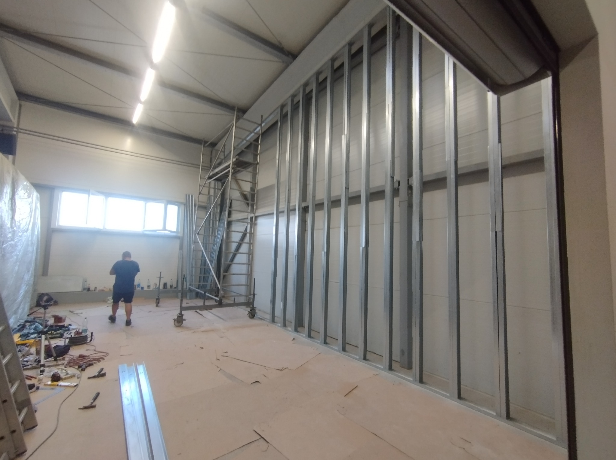 Metalowa konstrukcja szkieletowa ścianki działowej w hali, widoczne profile pionowe i poziome, w tle rusztowanie i pracownik w oddali.