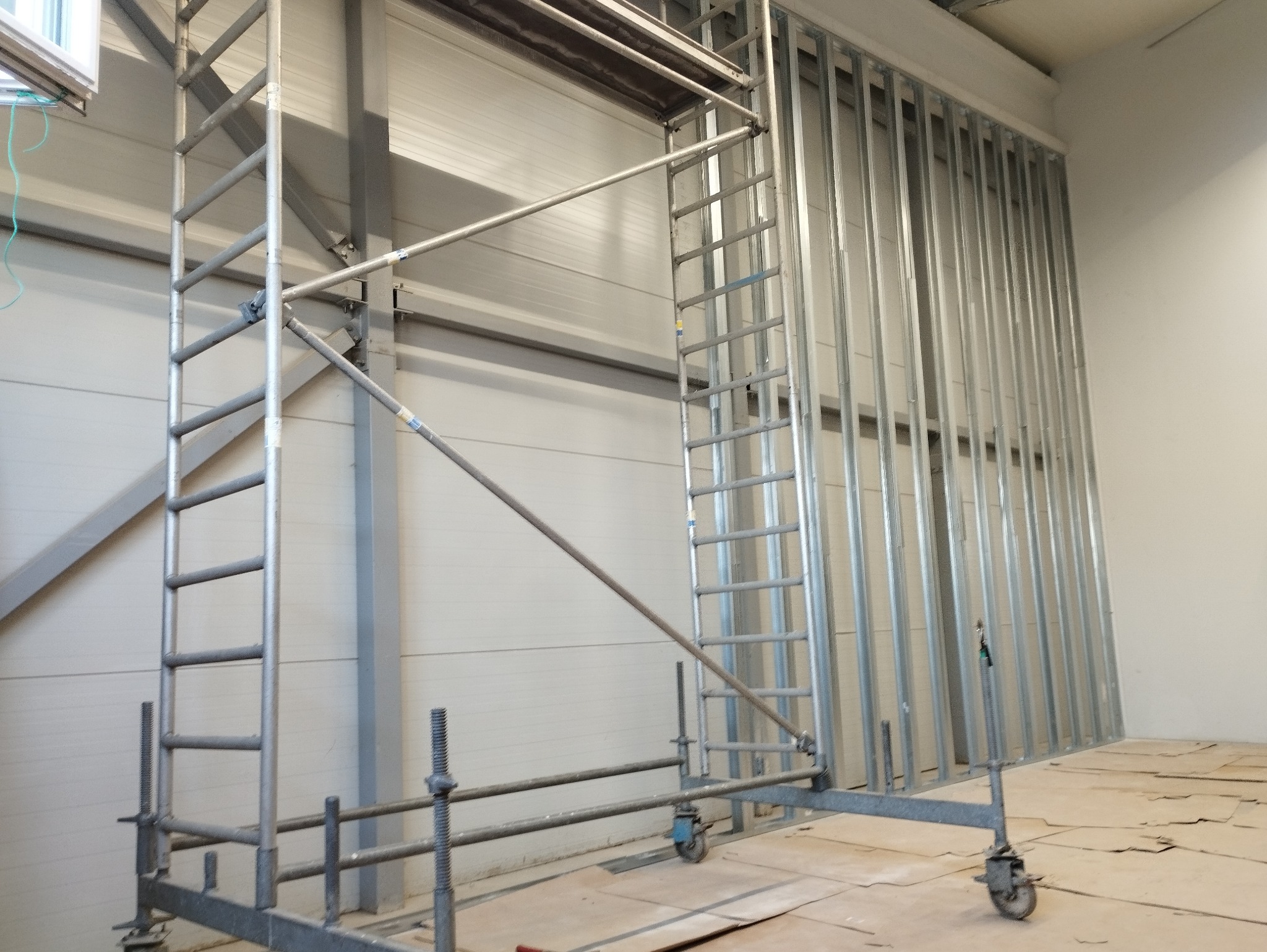 Stelaż z metalowych profili na podłodze zabezpieczonej kartonem, widoczne rusztowanie aluminiowe z regulowanymi podpórkami.