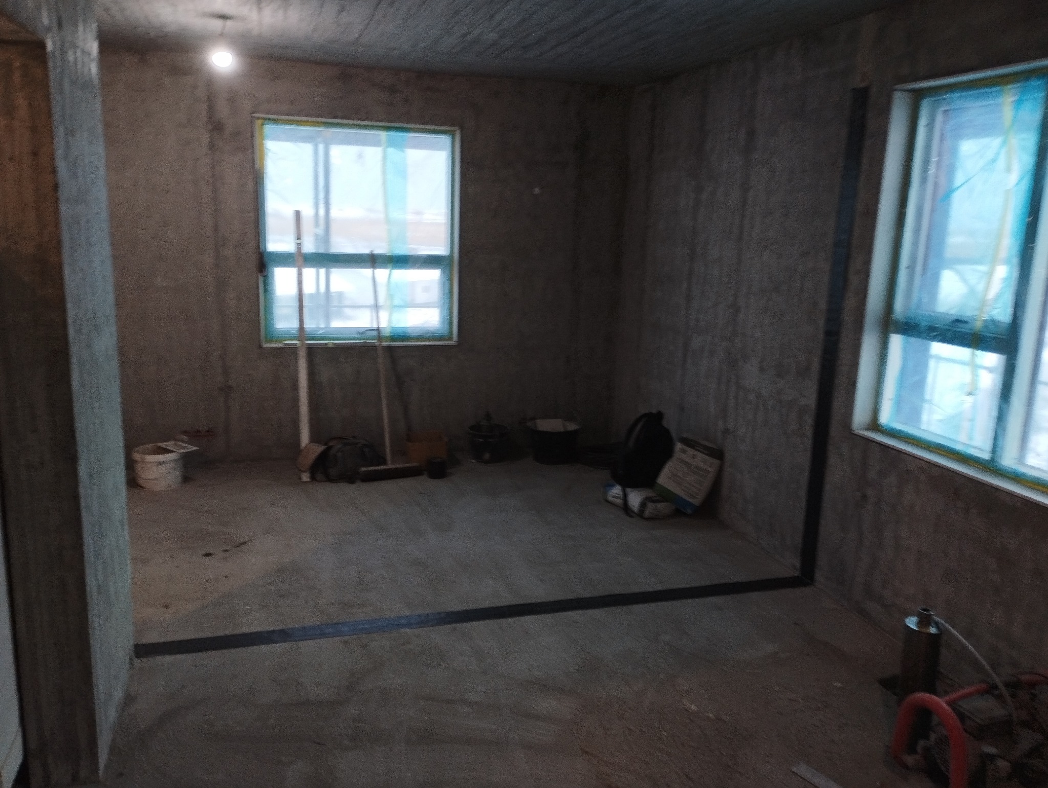 Surowy stan wnętrza domu w trakcie budowy: ściany z betonu komórkowego, okna zabezpieczone folią, widoczne narzędzia i materiały budowlane, brak wykończenia podłogi.