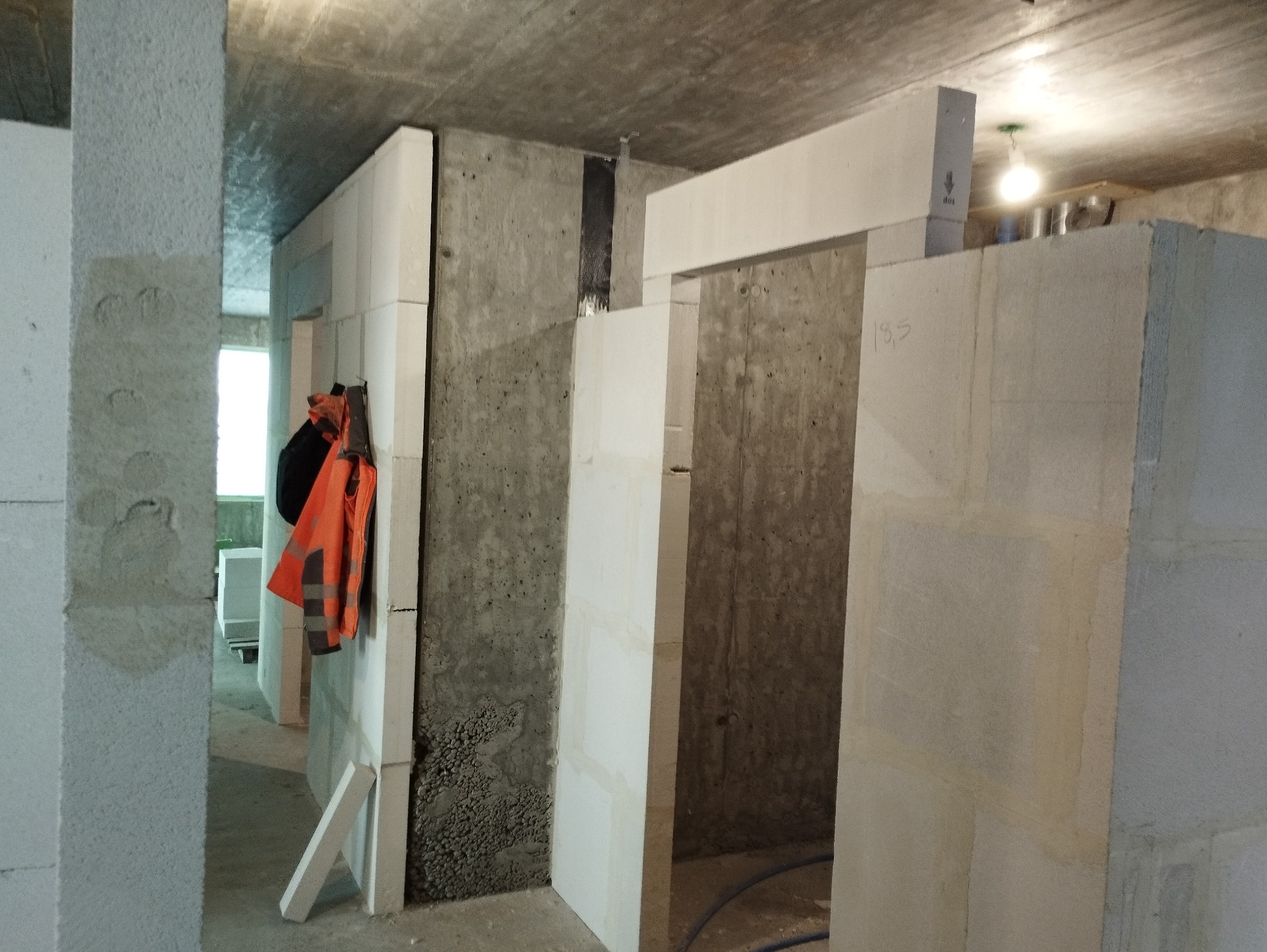 Wnętrze w trakcie budowy z surowymi ścianami z betonu komórkowego, widoczna pomarańczowa kamizelka odblaskowa i czarny plecak powieszone na ścianie, fragment instalacji elektrycznej na suficie.