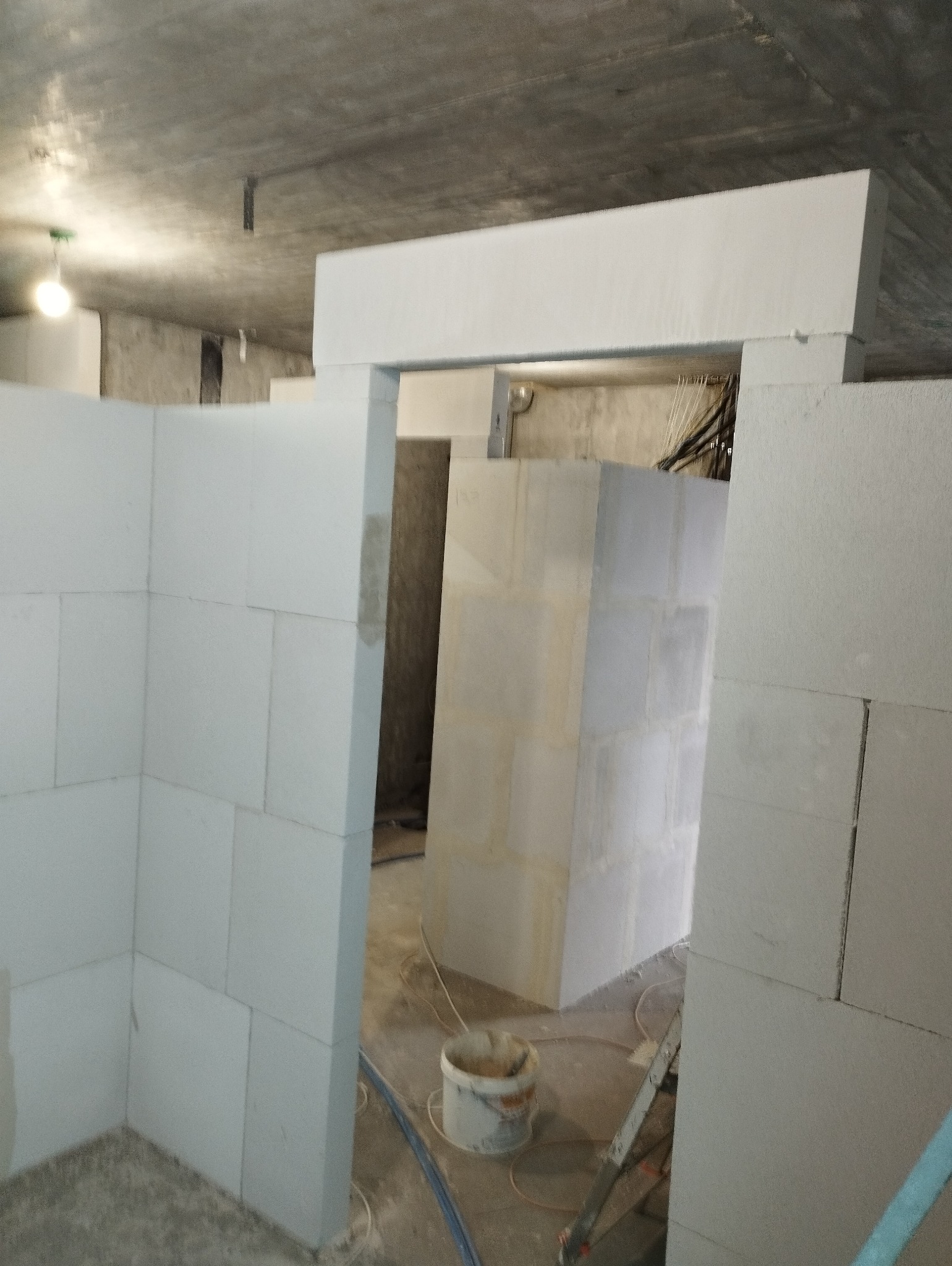 Wnętrze w trakcie budowy z białych bloczków, widoczny surowy betonowy sufit, wiadro z zaprawą i drabina w tle.