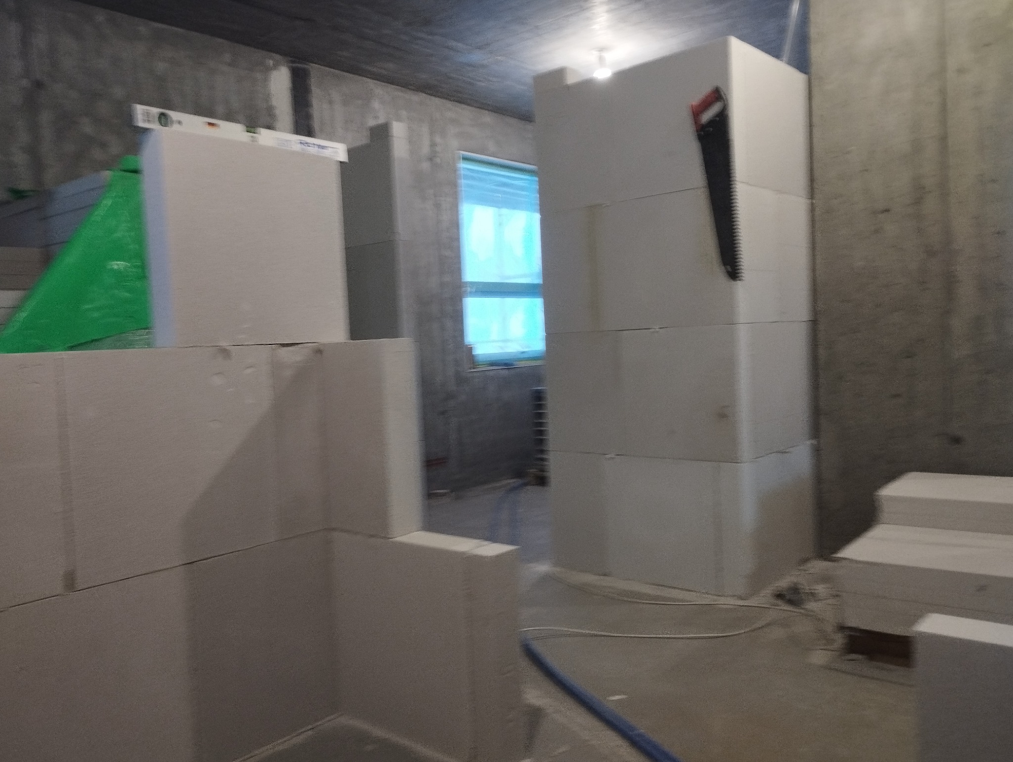 Wznoszenie ścian z białych bloczków, widoczna piła ręczna na ścianie, poziomica i folia ochronna, surowy stan wykończenia wnętrza.